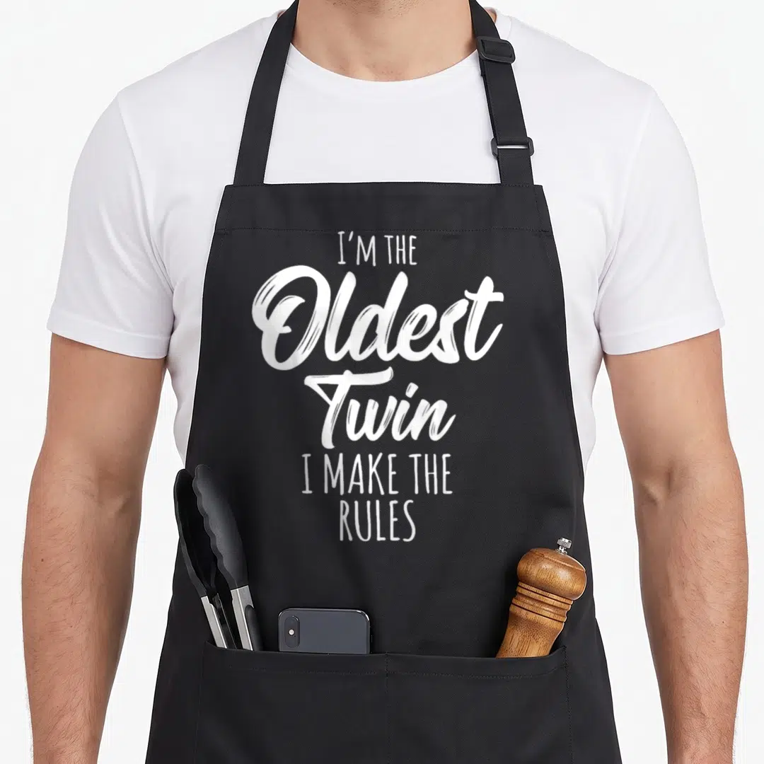 Sibling oldest birthday apron - White stacked text reading I'M THE Oldest Twin I MAKE THE RULES — shown on a black cotton canvas apron against a white background