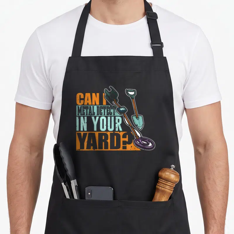 Detector metal detecting apron - Stacked text reading CAN I METAL DETECT IN YOUR YARD next to crossed detector — shown on a black cotton canvas apron against a white background