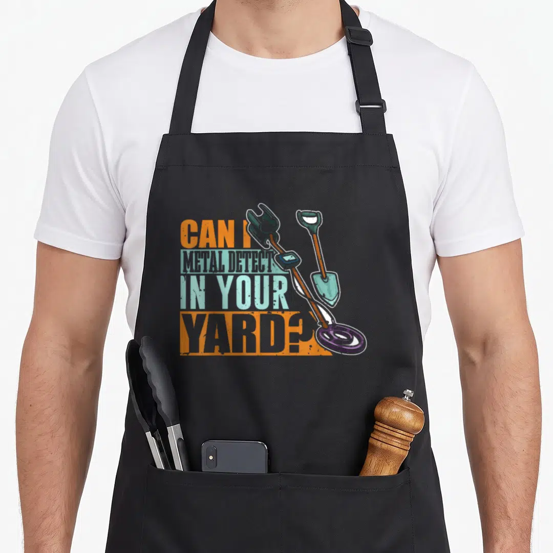 Detector metal detecting apron - Stacked text reading CAN I METAL DETECT IN YOUR YARD next to crossed detector — shown on a black cotton canvas apron against a white background