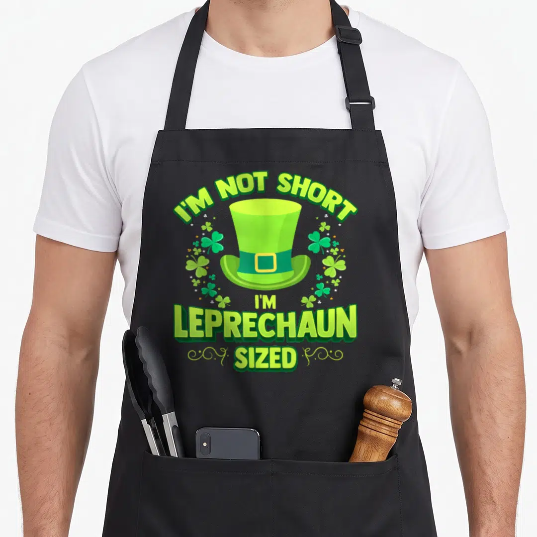 Leprechaun irish aprons - Green leprechaun hat with I'M NOT SHORT I'M LEPRECHAUN SIZED text and shamrock accents — shown on a black cotton canvas apron against a white background
