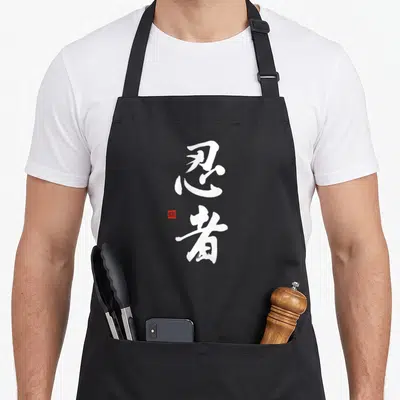 Ninja calligraphy chinese apron - Two vertical white kanji characters with a small red square seal stamp on the left — shown on a black cotton canvas apron against a white background