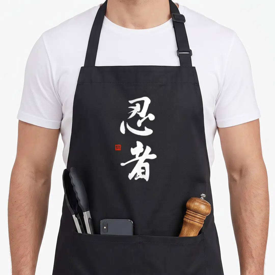 Ninja calligraphy chinese apron - Two vertical white kanji characters with a small red square seal stamp on the left — shown on a black cotton canvas apron against a white background