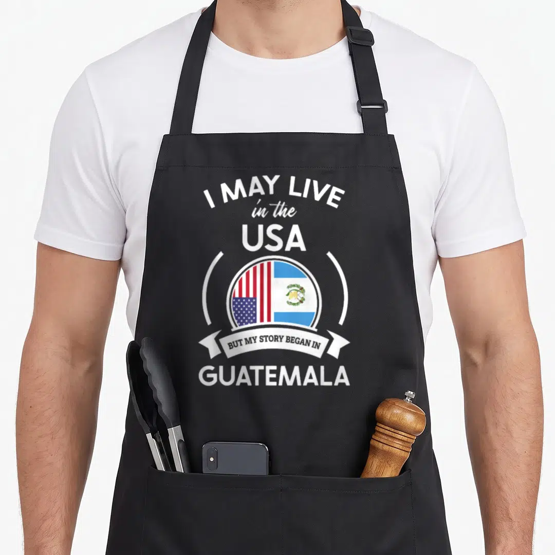 Guatemala story of the apron - Split US and Guatemala flags in circle with white I May Live in USA text — shown on a black cotton canvas apron against a white background