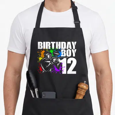 Paintball 12th birthday apron - Grey paintball player surrounded by red yellow green blue purple and orange paint — shown on a black cotton canvas apron against a white background