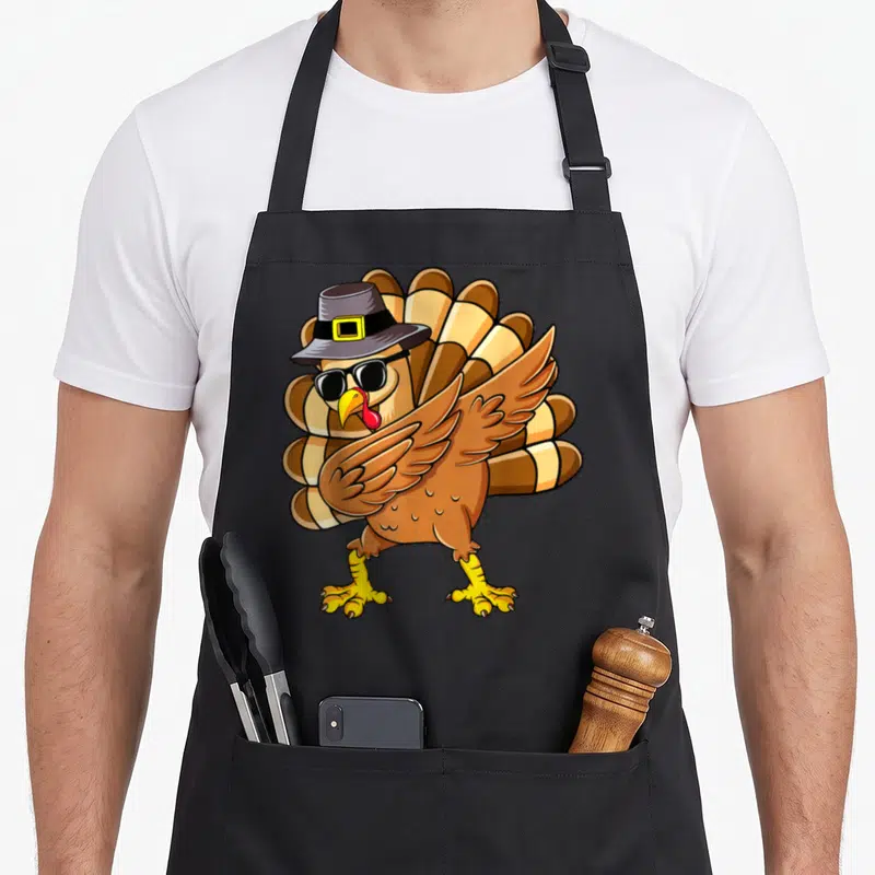 Dabbing turkey thanksgiving day aprons - Cartoon turkey wearing a grey pilgrim hat and black sunglasses performing — shown on a black cotton canvas apron against a white background