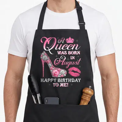 Congratulation queen apron - Pink high heel with diamonds, crown, lips, and A Queen Was Born in August text — shown on a black cotton canvas apron against a white background