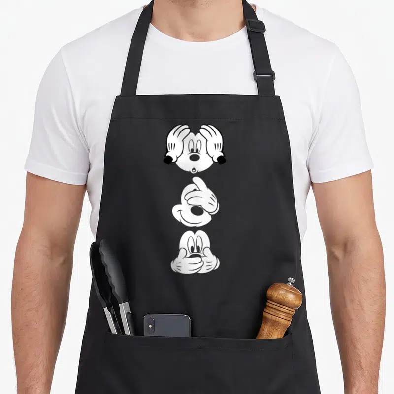 Mouse face apron - Three stacked Mickey Mouse heads with hands covering eyes, ears, and mouth in black and white — shown on a black cotton canvas apron against a white background