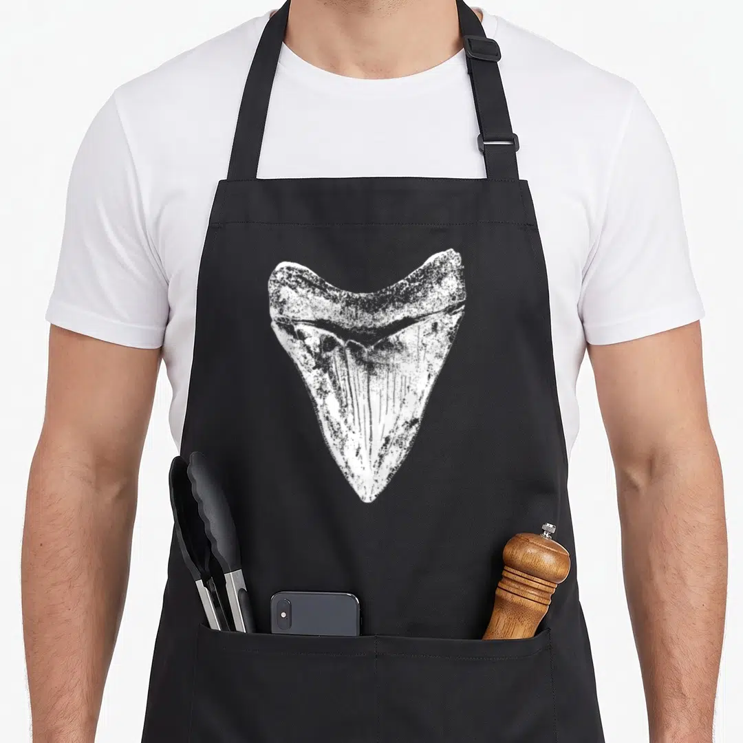 Megalodon science apron - White distressed shark tooth fossil print on black fabric — shown on a black cotton canvas apron against a white background