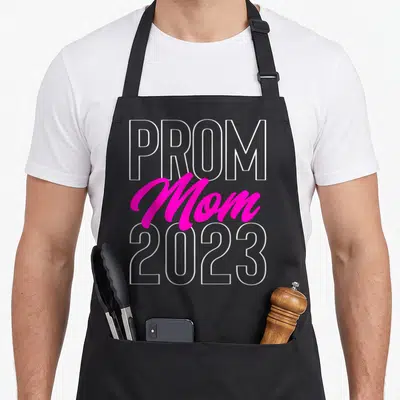 Prom apron or apron - Stacked PROM Mom 2023 text in white outline and pink script typography — shown on a black cotton canvas apron against a white background