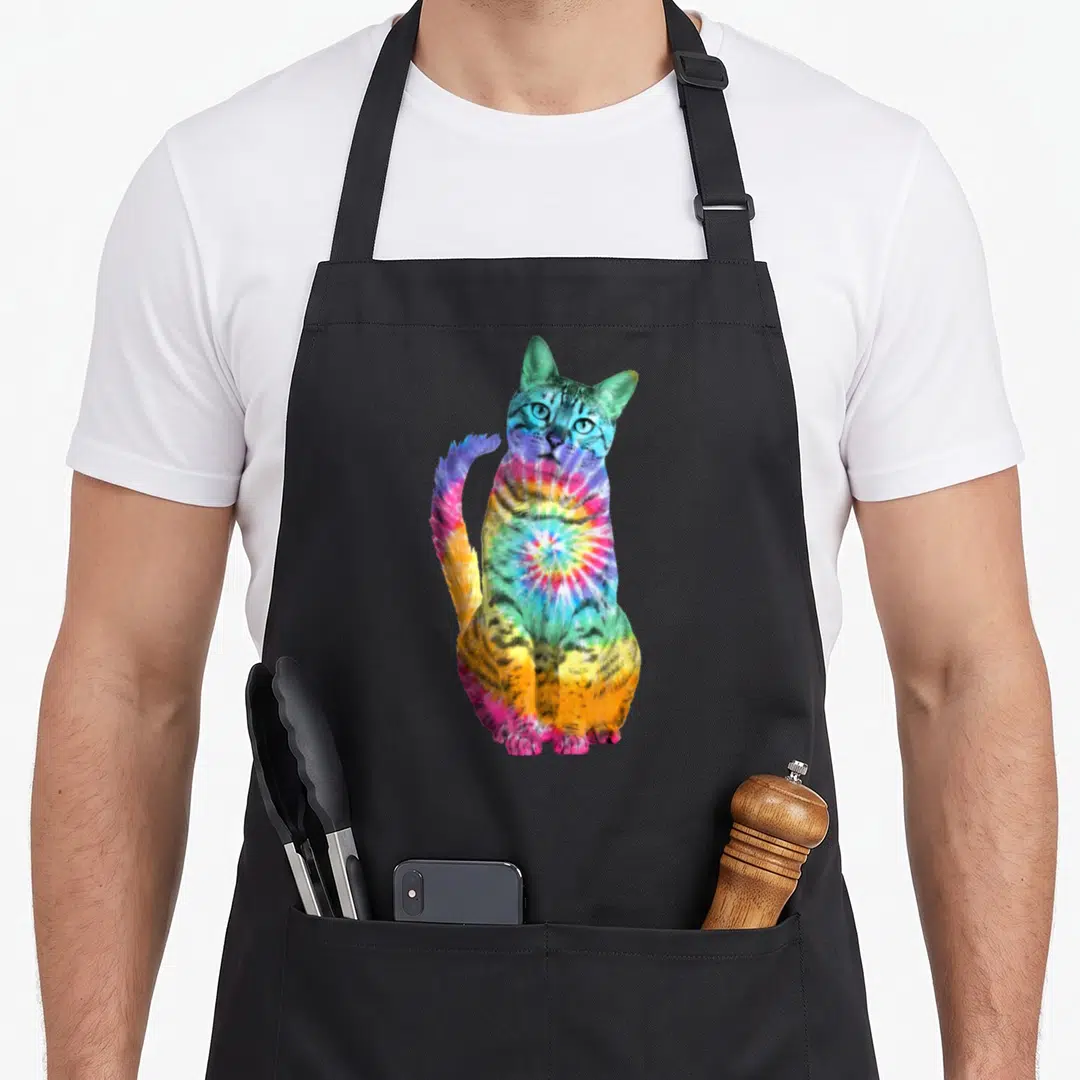 Cat tie dye apron - Sitting tabby cat rendered in rainbow tie-dye colors including blue, green, yellow, and pink — shown on a black cotton canvas apron against a white background
