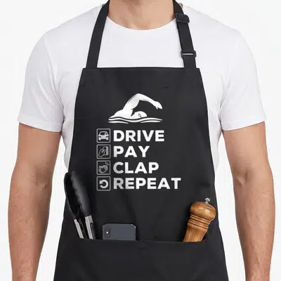 Drive swimming dad apron - White swimmer graphic above DRIVE PAY CLAP REPEAT text with icons — shown on a black cotton canvas apron against a white background