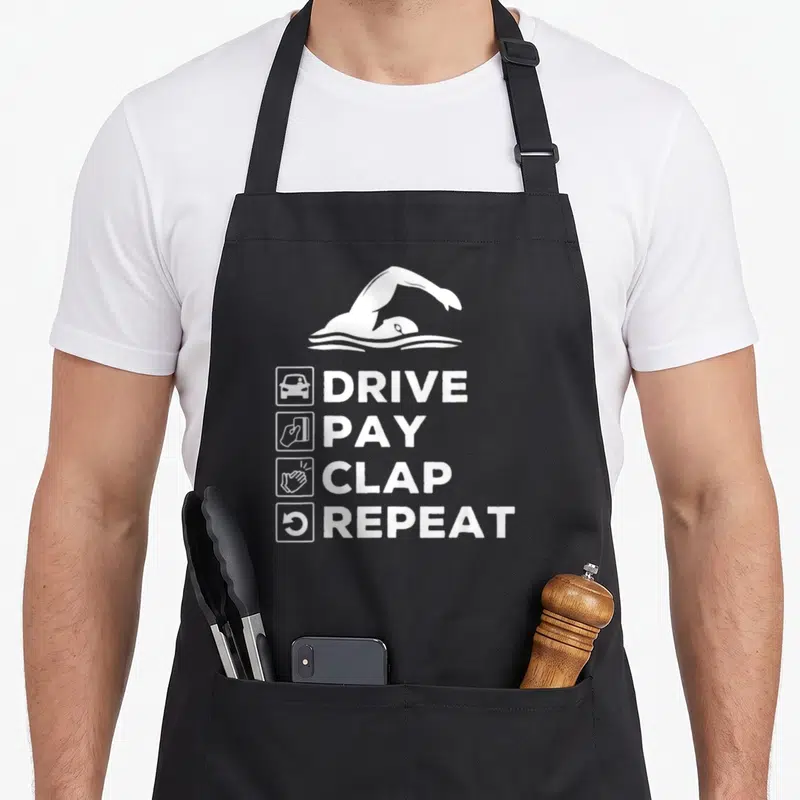Drive swimming dad apron - White swimmer graphic above DRIVE PAY CLAP REPEAT text with icons — shown on a black cotton canvas apron against a white background