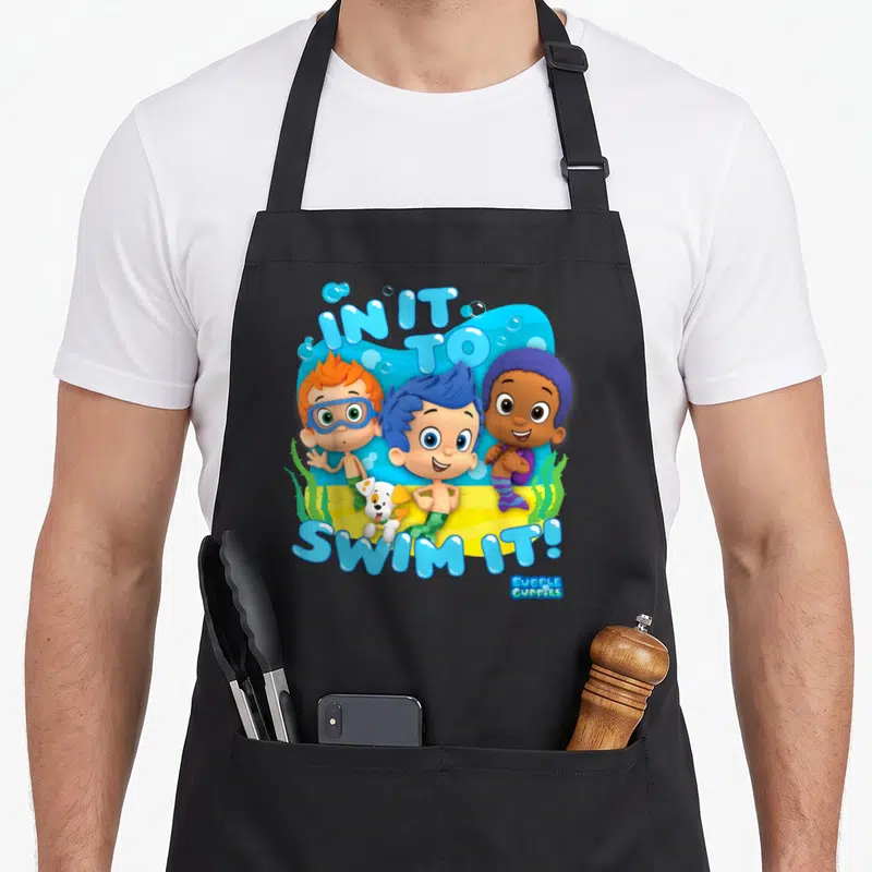 Bubble crazy guppy man apron - IN IT TO SWIM IT! text above three cartoon boys and dog on blue water and yellow sand — shown on a black cotton canvas apron against a white background