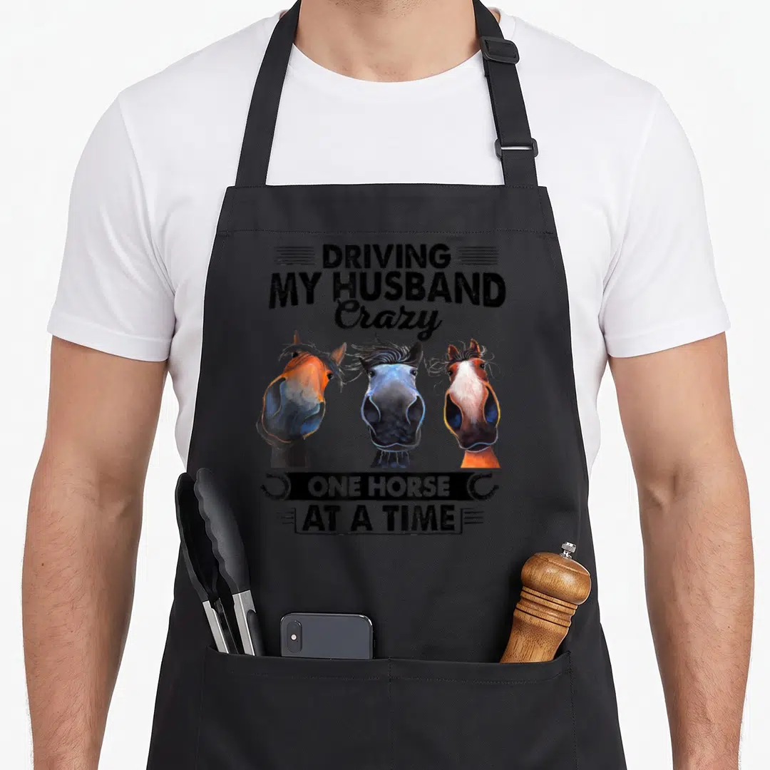 Horse driving aprons - Three horse heads with text Driving My Husband Crazy One Horse At A Time flanked by horseshoes — shown on a black cotton canvas apron against a white background