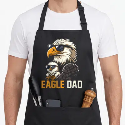 Eagle baby father day apron - Cartoon eagle and chick wearing sunglasses with EAGLE DAD text in orange and white — shown on a black cotton canvas apron against a white background