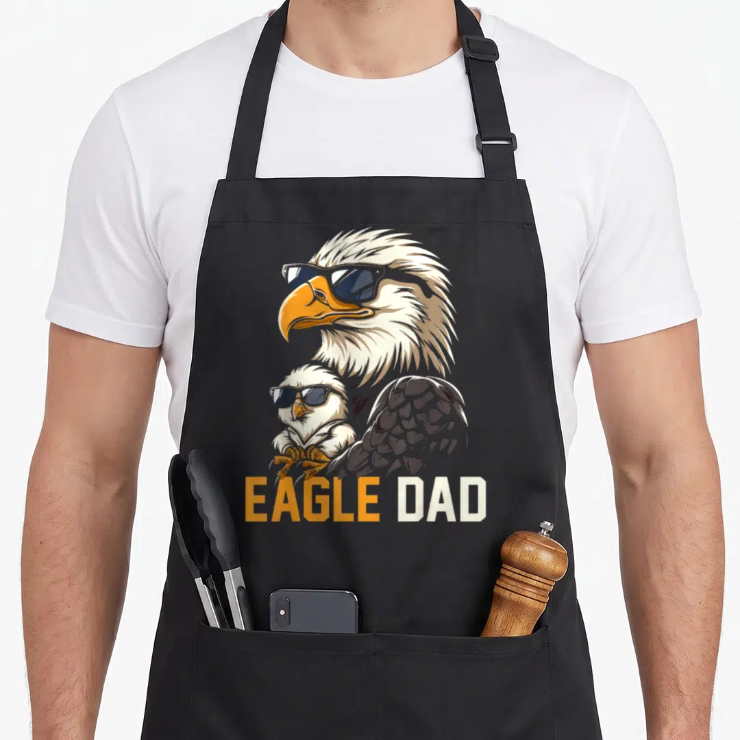 Eagle baby father day apron - Cartoon eagle and chick wearing sunglasses with EAGLE DAD text in orange and white — shown on a black cotton canvas apron against a white background