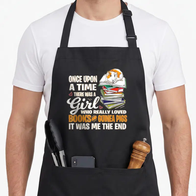 Loved pig lady apron - Cartoon guinea pig on books stack with quote about loving reading and pets — shown on a black cotton canvas apron against a white background