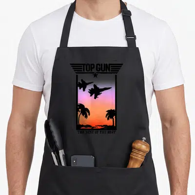 Gun tree apron - Two fighter jets flying over sunset beach with palm trees under TOP GUN text — shown on a black cotton canvas apron against a white background