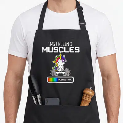 Installing muscle apron - Cartoon unicorn lifting grey barbell with rainbow mane and pixelated text — shown on a black cotton canvas apron against a white background