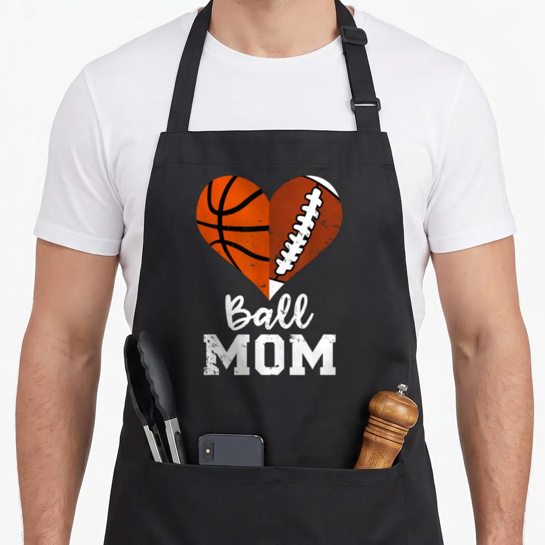 Basketball football apron - Split heart graphic with orange basketball and brown football above Ball MOM text — shown on a black cotton canvas apron against a white background