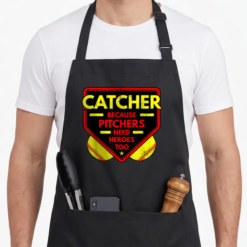 Pitcher softball apron - Catcher Because Pitchers Need Heroes Too text on red shield with two yellow baseballs — shown on a black cotton canvas apron against a white background