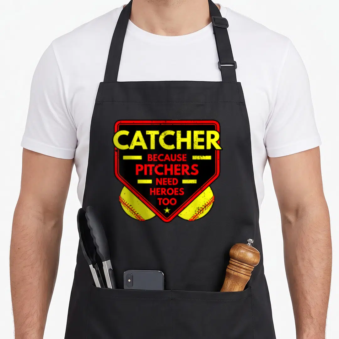 Pitcher softball apron - Catcher Because Pitchers Need Heroes Too text on red shield with two yellow baseballs — shown on a black cotton canvas apron against a white background