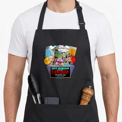 Jeff tacoma apron - Cartoon puppets behind grey barrier reading Jeff Dunham Still Not Canceled Tacoma WA March 26 2023 — shown on a black cotton canvas apron against a white background