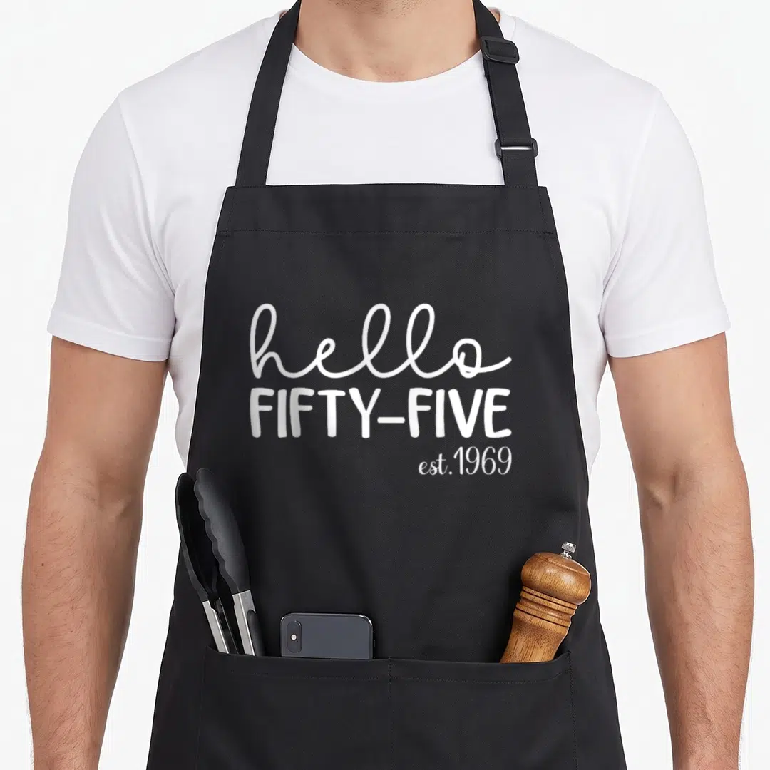Five est birthday apron - White text reading hello FIFTY-FIVE est.1969 on grey background — shown on a black cotton canvas apron against a white background