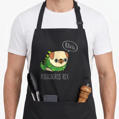 Pug cheap halloween aprons - Cartoon pug in green spotted dinosaur costume with PUGASAURUS REX text and speech bubble — shown on a black cotton canvas apron against a white background