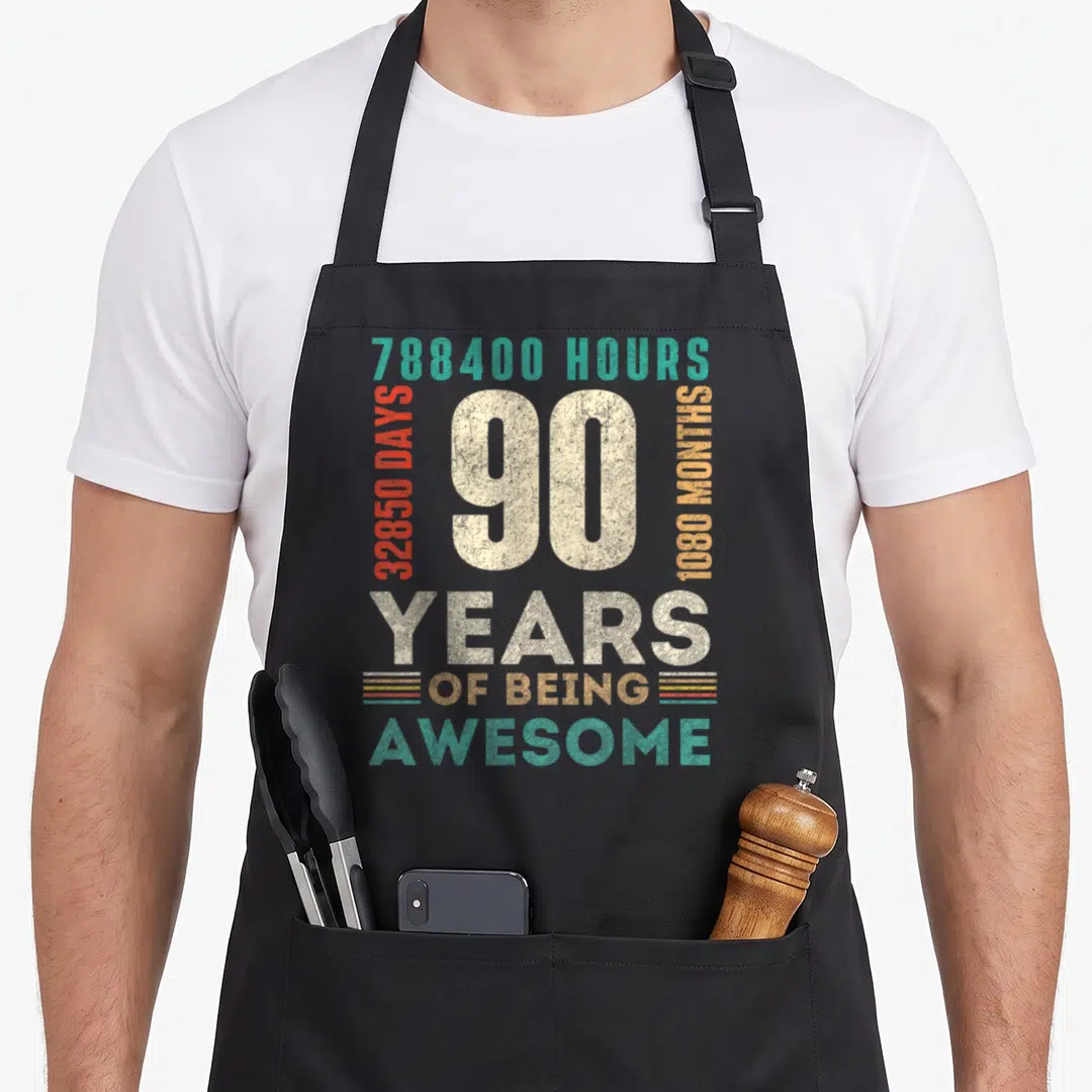 Hour birthday apron - Distressed 90 years of being awesome text with hours days months stats in beige teal red orange — shown on a black cotton canvas apron against a white background