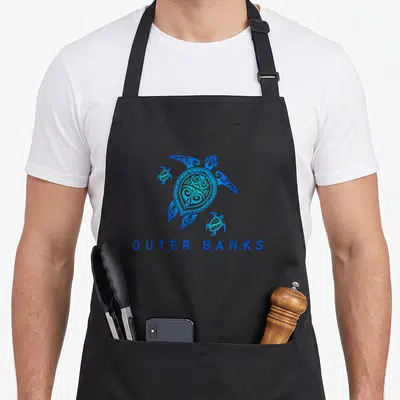 North outer banks apron - Tribal sea turtles above OUTER BANKS text in blue on gray background — shown on a black cotton canvas apron against a white background