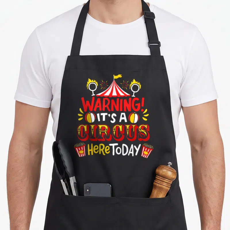 Funny circus apron - Red and white circus tent with WARNING IT'S A CIRCUS text, fire rings and popcorn boxes in cartoon — shown on a black cotton canvas apron against a white background