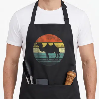 Camel zoo animal apron - Grey camel silhouette inside a retro circle of orange, yellow, teal, and grey horizontal — shown on a black cotton canvas apron against a white background