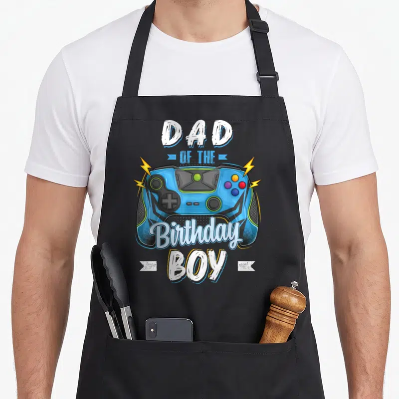 Video family aprons - Blue game controller with yellow bolts and white text Dad of the Birthday Boy — shown on a black cotton canvas apron against a white background