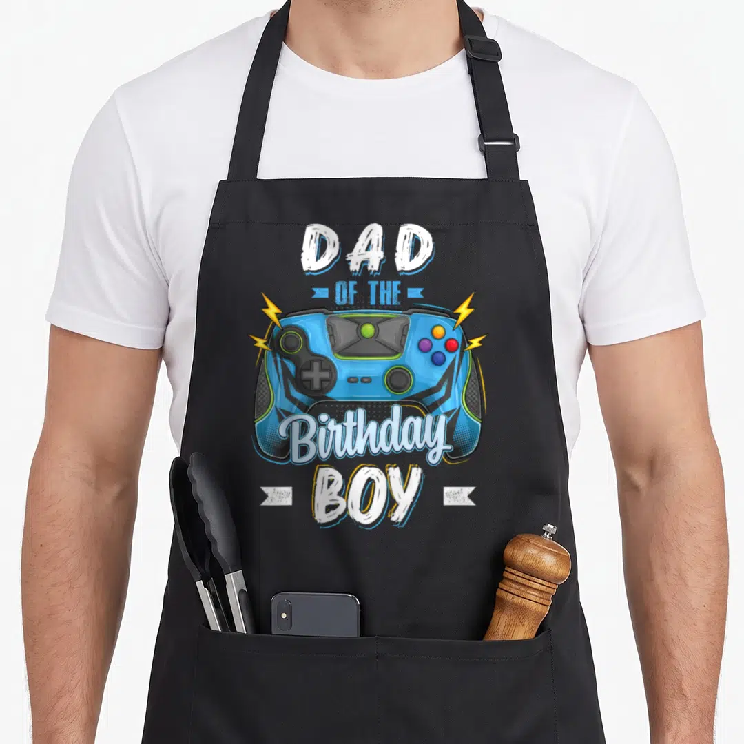 Video family aprons - Blue game controller with yellow bolts and white text Dad of the Birthday Boy — shown on a black cotton canvas apron against a white background