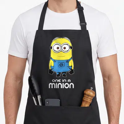 Despicable bobsled apron - Yellow minion in blue overalls above white text ONE IN A MINION — shown on a black cotton canvas apron against a white background