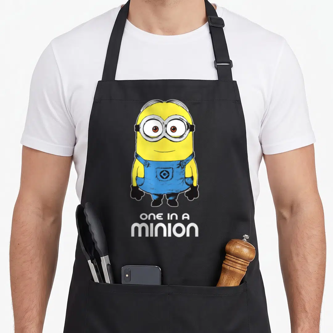 Despicable bobsled apron - Yellow minion in blue overalls above white text ONE IN A MINION — shown on a black cotton canvas apron against a white background