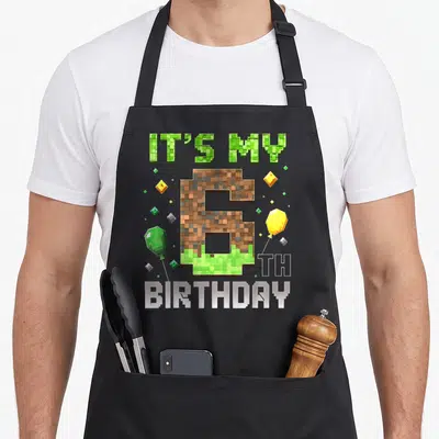 6th birthday video game apron - Pixelated number 6 with dirt texture, gem balloons, and IT'S MY 6 TH BIRTHDAY text — shown on a black cotton canvas apron against a white background