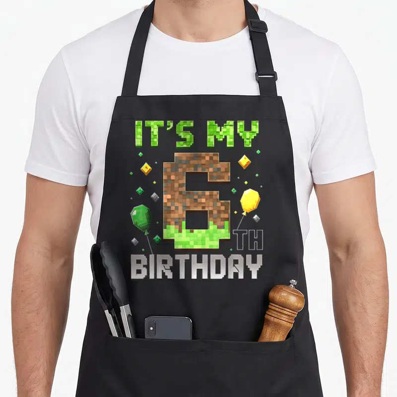 6th birthday video game apron - Pixelated number 6 with dirt texture, gem balloons, and IT'S MY 6 TH BIRTHDAY text — shown on a black cotton canvas apron against a white background