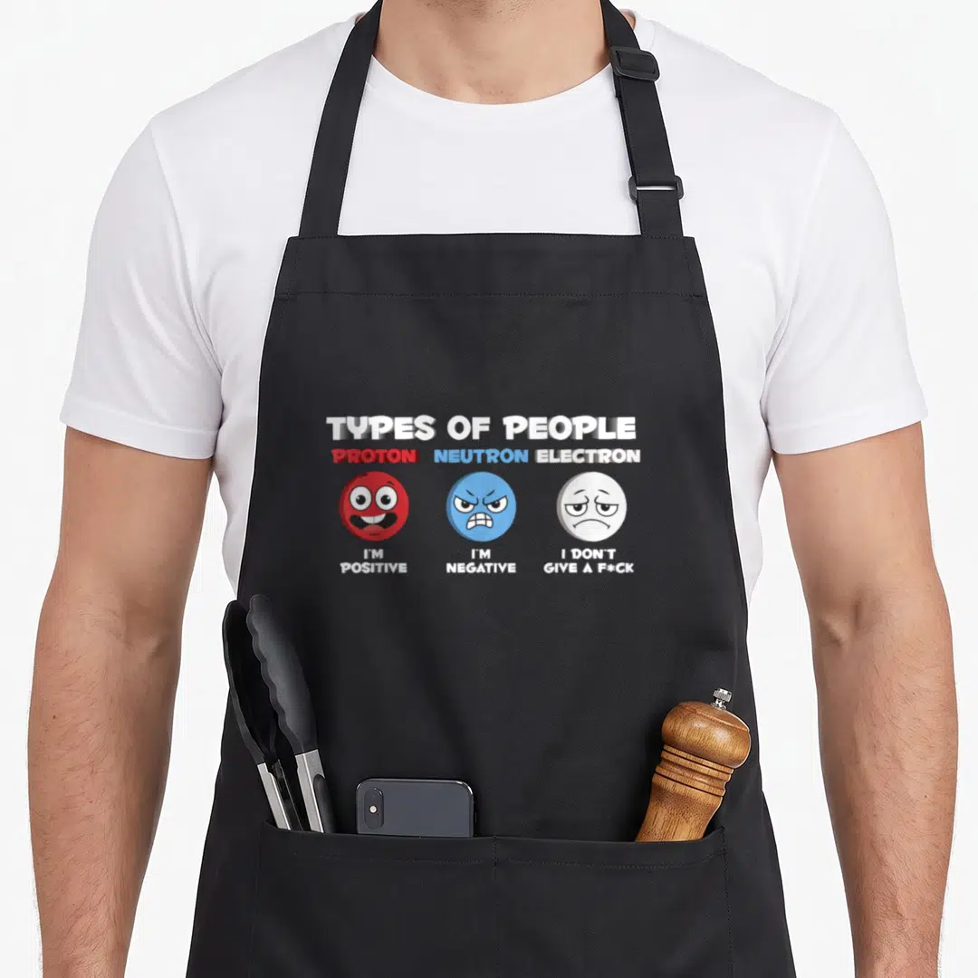 Physicist funny nerd apron - Three cartoon faces in red, blue, white labeled proton, neutron, electron with personality — shown on a black cotton canvas apron against a white background