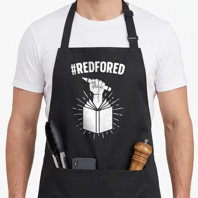 Strike teacher apron - White distressed #REDFORED text above a fist holding a broken pencil emerging from an open book — shown on a black cotton canvas apron against a white background