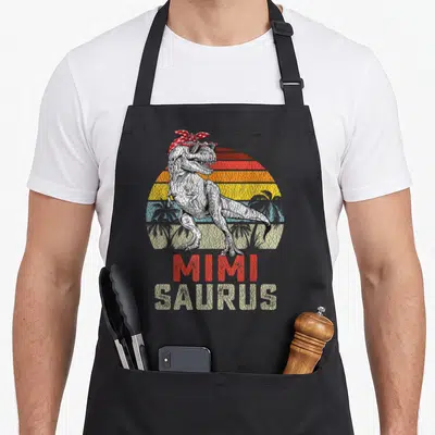 Mimisaurus dinosaur apron - Retro Tyrannosaurus Rex with sunglasses and bandana over striped sun with MIMI SAURUS text — shown on a black cotton canvas apron against a white background