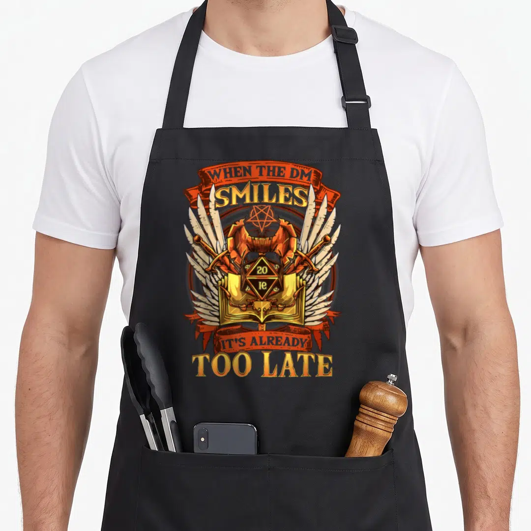 Smile salute apron - Dungeon master kit with d20 showing 20, wings, and sarcasm text in orange and gold — shown on a black cotton canvas apron against a white background