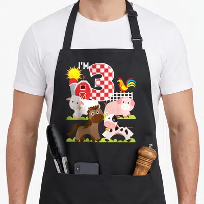Barnyard farm apron - I'M 3 text with checkered number, barn, rooster, cow, pig, horse, sheep, sun — shown on a black cotton canvas apron against a white background