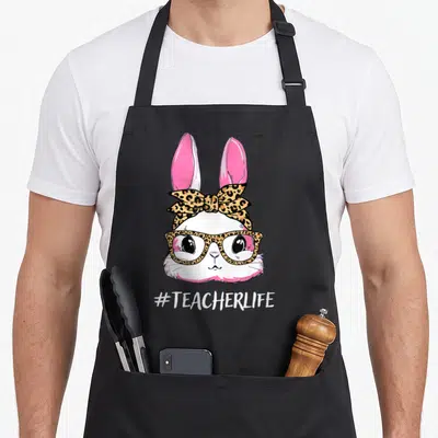 Hoppy teacher easter bunny aprons - Cartoon white bunny with pink ears wearing leopard glasses and bow above — shown on a black cotton canvas apron against a white background