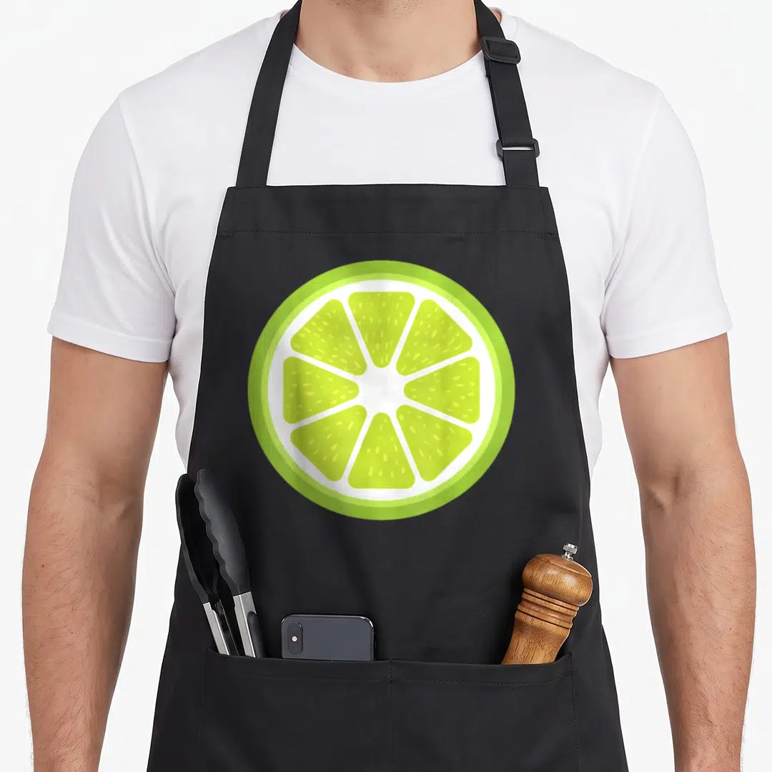 Sliced fruit apron - Circular lime slice in green and white with segmented pulp and rind — shown on a black cotton canvas apron against a white background