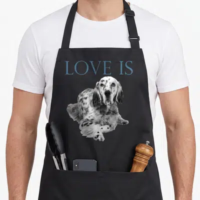 Funny setter english apron - Blue LOVE IS text above a black and white photo of a resting spotted dog — shown on a black cotton canvas apron against a white background