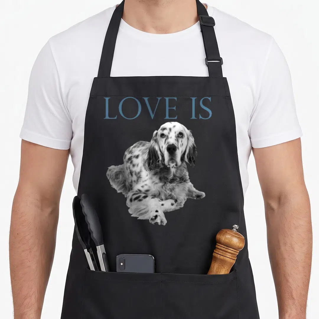 Funny setter english apron - Blue LOVE IS text above a black and white photo of a resting spotted dog — shown on a black cotton canvas apron against a white background