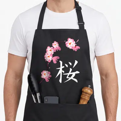 Festival cherry apron - Pink and white cherry blossom branch curving beside a large white kanji character on black — shown on a black cotton canvas apron against a white background