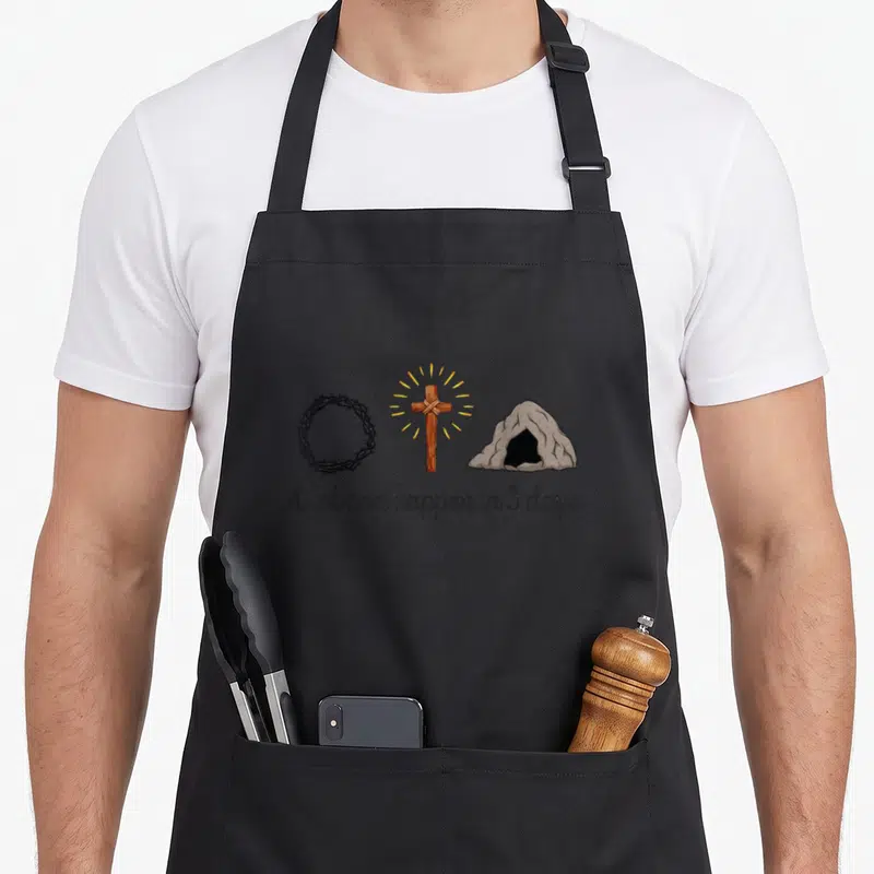 Happen christian easter apron - Crown of thorns, glowing wooden cross, and stone tomb above script text on grey — shown on a black cotton canvas apron against a white background